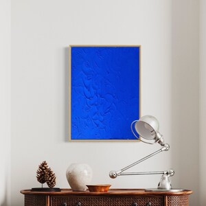 May include: A blue abstract painting with a textured surface in a gold frame. The painting is hanging on a white wall above a wooden table with a lamp and decorative objects.