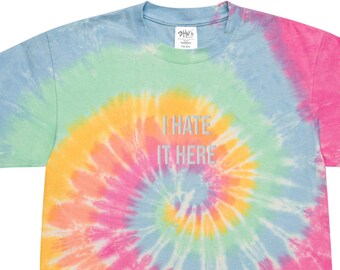 May include: A tie-dye t-shirt with a spiral pattern in shades of blue, green, yellow, pink, and orange. The shirt features the text "I HATE IT HERE" in silver lettering. The shirt is a short-sleeved crew neck.