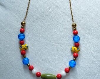 May include: A colorful necklace with a gold chain. The necklace features red, blue, and white beads, along with two yellow fish-shaped beads. A green oval bead is centered at the bottom.