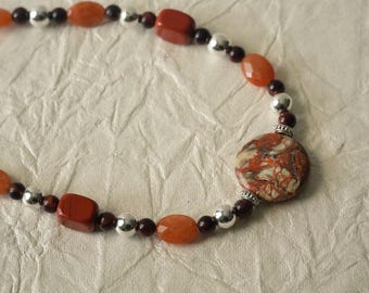 May include: A beaded necklace featuring a large, round, multi-colored patterned stone pendant as the focal point. The strand consists of alternating orange oval beads, dark red square beads, small dark brown round beads, and shiny silver spheres.