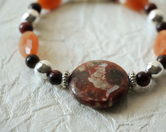 May include: A beaded bracelet featuring a large, round, mottled brown and red jasper focal bead flanked by silver-toned spacers. The design includes alternating dark brown round beads, faceted orange oval beads, and polished silver-toned spheres.