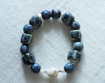 May include: A handmade bracelet featuring a mix of blue and white beads. The bracelet includes square and round beads in shades of blue with a speckled design, interspersed with smaller white beads. The bracelet is displayed on a white surface.
