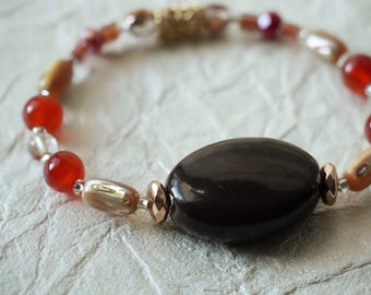 May include: A beaded bracelet featuring a large, smooth, dark brown oval center bead flanked by smaller red spherical beads, iridescent tan cylindrical beads, and metallic accents, all strung together on a flexible cord.