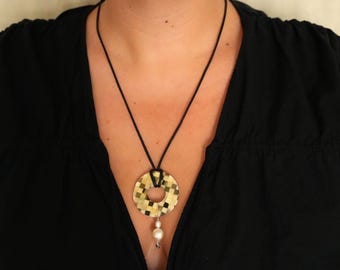 May include: A necklace featuring a circular pendant with a green and gold checkered pattern suspended from a thin black cord. A single white pearl-like bead hangs from the bottom of the circular pendant, adding a subtle accent to the design.
