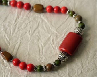 May include: A beaded necklace featuring a large rectangular red bead flanked by silver-toned ribbed spacers. The strand alternates between round red beads, round green and brown mottled beads, and brown rectangular beads with small silver-toned accents.