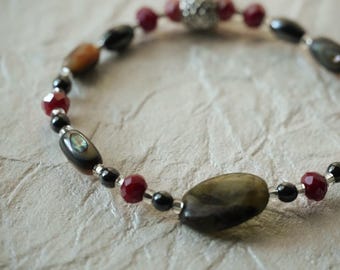 May include: A beaded bracelet featuring a mix of dark brown and tan polished stones, faceted deep red beads, small black spherical beads, and a single textured silver-toned metal bead, all strung together on a flexible cord.