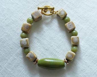 May include: A handmade bracelet featuring a toggle clasp and a mix of square and round beads. The square beads are tan with white accents, while the round beads are a light green. A larger, olive-green oval bead is centered.