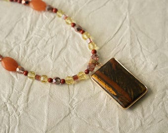 May include: A necklace featuring a rectangular tiger's eye pendant set in a gold-toned frame. The chain consists of a variety of beads, including orange oval stones, small red spheres, faceted yellow crystals, and translucent purple-tinted beads.