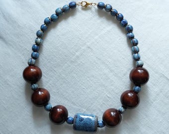May include: A handcrafted necklace featuring a mix of dark brown wooden beads, speckled blue ceramic beads, and a rectangular blue ceramic bead. The necklace is strung on a gold-colored chain and clasp, creating a unique and earthy accessory.