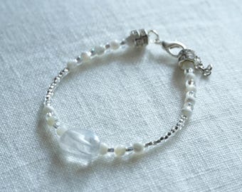 May include: A delicate bracelet featuring a clear, oblong gemstone as a focal point, complemented by small white beads and silver-toned accents. The bracelet is strung on a fine, silver chain and has a secure clasp.