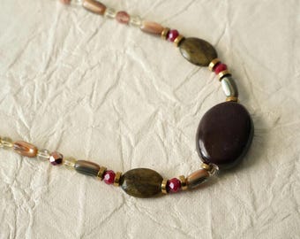 May include: A beaded necklace featuring a large, smooth, dark brown oval focal bead, accented with smaller olive green oval beads, faceted red glass beads, metallic gold-tone spacers, and elongated iridescent brown cylindrical beads.