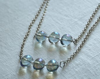 May include: Two delicate necklaces with silver chains. Each necklace features three iridescent, clear glass beads. The necklaces are displayed on a white, textured surface, creating a simple and elegant aesthetic.