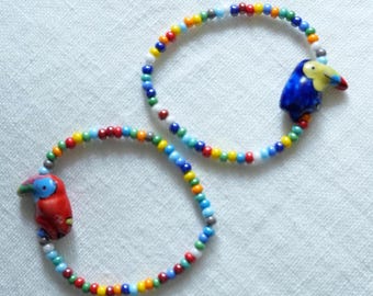 May include: Two beaded bracelets with colorful beads in a rainbow pattern. Each bracelet features a ceramic toucan charm. One toucan is red, and the other is blue. The bracelets are on a white surface.