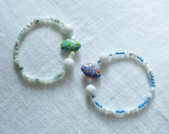 May include: Two beaded bracelets with fish charms. One bracelet features a turquoise and yellow fish charm, while the other has an orange and blue fish. The bracelets are made with white and blue beads.