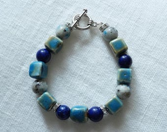 May include: A beaded bracelet featuring a mix of blue, turquoise, and gray beads. The bracelet includes square and round beads, with a silver toggle clasp. The bracelet is approximately 7 inches in length.
