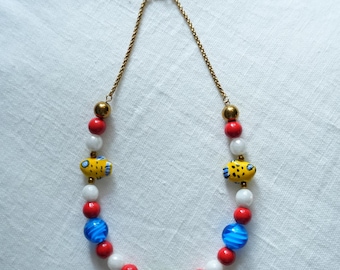 May include: A colorful beaded necklace featuring a gold chain and a variety of beads. The necklace includes red, white, and blue beads, along with two yellow fish-shaped beads. A green oval bead is also present.