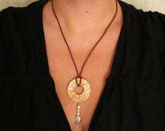 May include: A necklace featuring a brown cord and a circular, hammered-texture pendant in a light beige tone. A small vertical drop hangs from the center of the pendant, adorned with a series of small, light-colored beads and a heart-shaped charm.