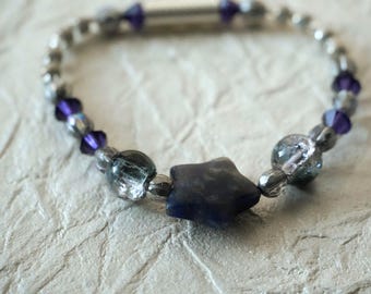 May include: A beaded bracelet featuring a central dark blue star-shaped bead, flanked by two clear crackle-textured glass beads, and accented with alternating small metallic silver beads and deep purple faceted crystal bicone beads throughout.