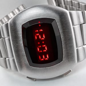 Può includere: Un orologio da polso digitale in metallo color argento spazzolato con cassa rettangolare arrotondata e cinturino a maglie metalliche abbinato. Il quadrante presenta uno schermo nero con numeri LED rosso vivo che indicano le 12:03.