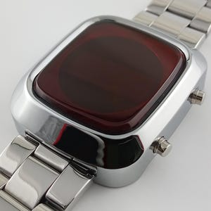 May include: A digital wristwatch featuring a polished silver-tone metal link band and a rectangular case with rounded corners. The watch face is covered by a dark red, translucent glass display panel, and two small silver buttons are on the side.