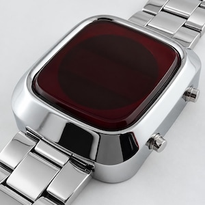 May include: A wristwatch with a polished silver-tone metal link band and a rectangular case. The watch face features a dark red, smooth, rounded glass surface with no visible markings or hands, and two small silver buttons on the right side.