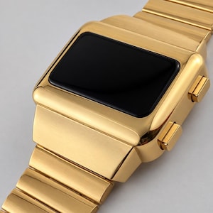 May include: A gold-toned digital watch featuring a rectangular case with a smooth, black, blank display screen. The timepiece includes a matching gold-toned metal link bracelet and two rectangular push buttons located on the side of the watch case.