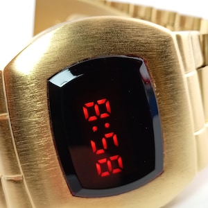 Può includere: Un orologio da polso digitale color oro con finitura in metallo spazzolato e cassa rettangolare. L'orologio presenta uno schermo nero che mostra l'ora in cifre LED rosso brillante. Include un cinturino a maglie in metallo color oro abbinato.