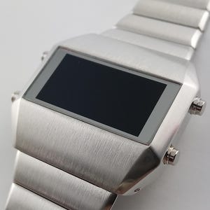 Könnte beinhalten: Eine silberfarbene Digitalarmbanduhr mit einem rechteckigen schwarzen Bildschirm und gebürsteter Metalloberfläche. Die Uhr hat ein segmentiertes Metallarmband und zwei Knöpfe an der Seite. Die Uhr ist im Retro-Stil.