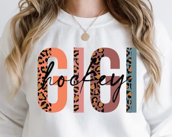 May include: White sweatshirt with the text "GIGI hockey" in a colorful design. The letters "GIGI" are in orange, brown, and leopard print, with the word "hockey" in a script font.