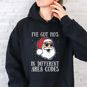 May include: Black hooded sweatshirt with the text "I've got ho's in different area codes" and a graphic of Santa Claus wearing sunglasses. The Santa graphic has a red hat, white beard, and a smiling face. The sweatshirt is a casual style.