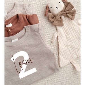Peut inclure: Un t-shirt gris avec un numéro "2" blanc et le mot "Leon" écrit en cursive. Le t-shirt est sur une surface texturée beige avec un jouet en peluche brun et beige.