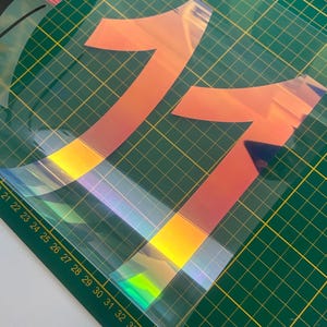 Op de afbeelding: Twee regenboogkleurige vinyl stickers in de vorm van het cijfer "1" op een groene snijmat. De stickers hebben een holografische afwerking met een regenboogeffect.