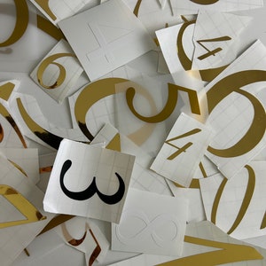 Op de afbeelding: Een verzameling gouden en zwarte nummerstickers op witte vellen papier. De nummers omvatten 1, 3, 4, 5, 6, 7, 8 en oneindig.