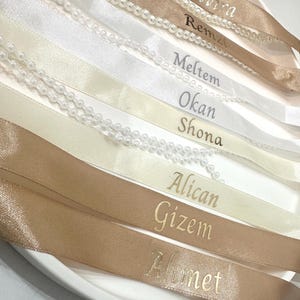 Könnte beinhalten: Nahaufnahme von mehreren Satinbändern in Weiß-, Creme- und Beigetönen, jedes mit einem Namen in gold- oder silberfarbener Schrift. Einige Bänder sind mit einer Reihe kleiner, weißer Perlen verziert.