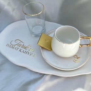 May include: White ceramic tray with gold trim, featuring a white coffee cup and saucer with gold trim, a glass of water with a gold rim, and a gold chocolate bar. The tray has the text "Tarik'in DAMAT KAHVESI" in gold lettering.