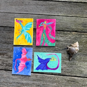 May include: Four rectangular art prints featuring colorful dragon illustrations. One shows a blue dragon on yellow, one a green-spotted creature on pink, one a pink dragon on blue, and one a blue dragon on light green. A single seashell is placed nearby.