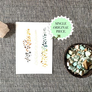 May include: A still-life composition featuring a white card with abstract designs in shades of brown, green, and yellow. A light-colored crystal and a small bowl filled with turquoise and brown stones are also present. The text "SINGLE ORIGINAL PIECE" is visible.