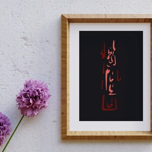 Puede incluir: Una impresión artística enmarcada con caligrafía roja sobre fondo negro. La obra de arte está encerrada en un marco de madera marrón claro. Dos flores moradas se colocan a la izquierda del marco contra una pared blanca texturizada.