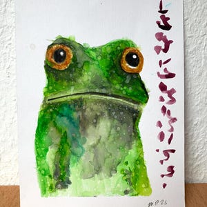 May include: Watercolor painting of a green frog with large orange eyes. The frog is centered on a white background. Japanese text is written vertically on the right side of the painting. The artist's signature is in the bottom right corner.