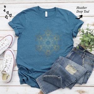 May include: A heather deep teal t-shirt with a gold geometric design. The shirt is laid flat on a white wooden surface with a pair of white sneakers and a pair of blue jeans.