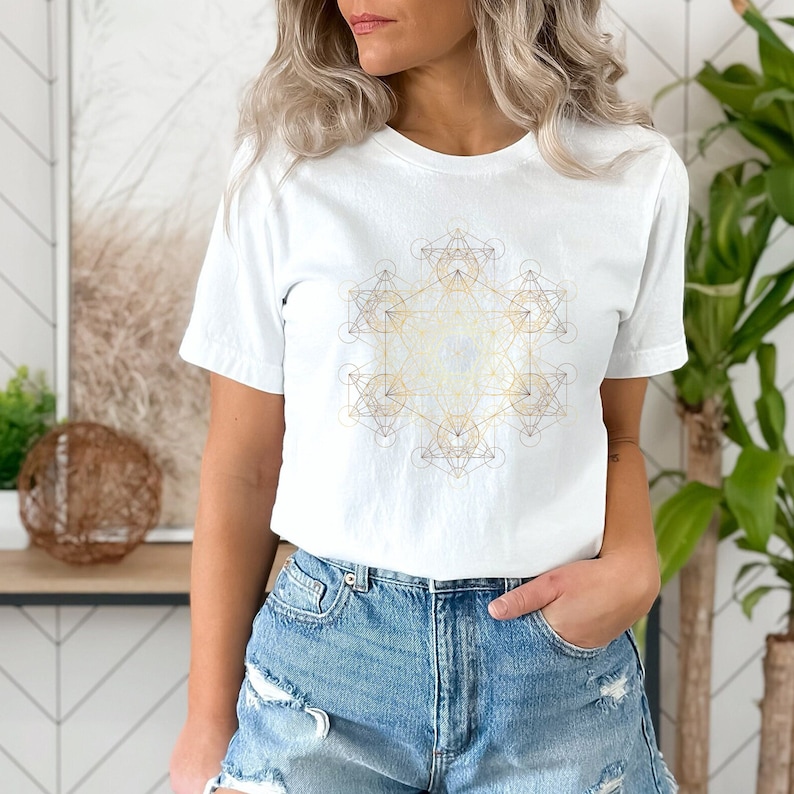 May include: White t-shirt with a gold geometric design on the front. The design is a complex pattern of overlapping circles, triangles, and squares.