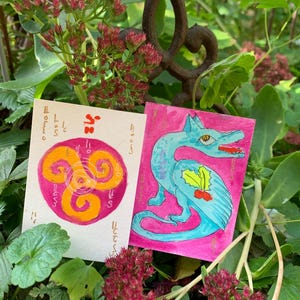 May include: Two rectangular art cards featuring hand-drawn illustrations. The left card displays an orange triskelion symbol on a pink circular background with red script. The right card shows a blue dragon with a holly leaf on a pink background.