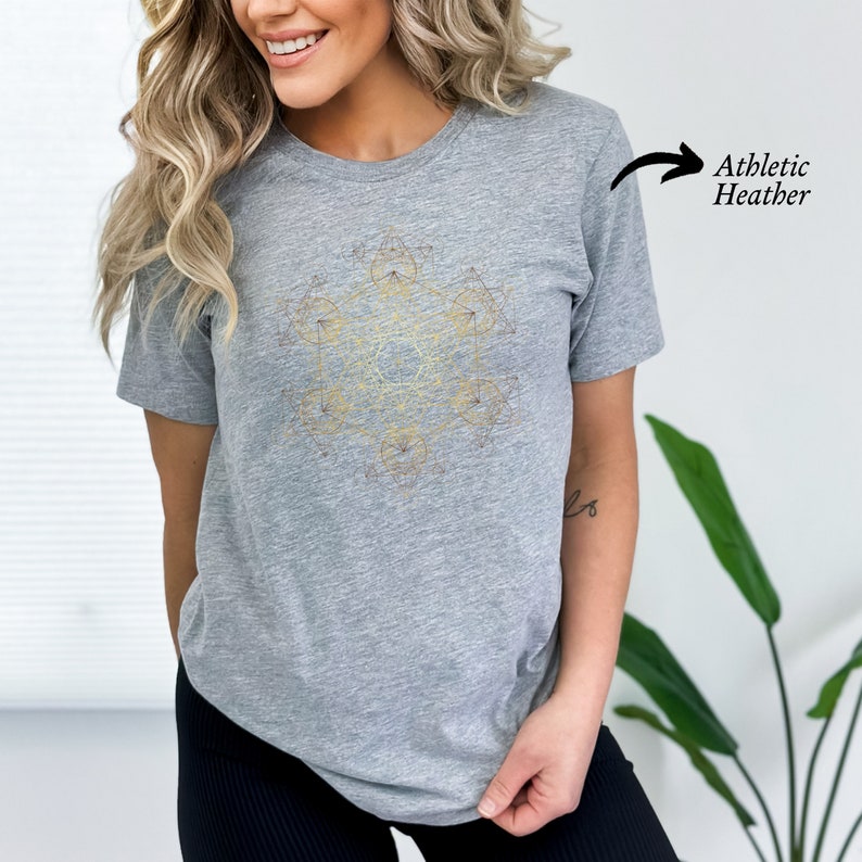 May include: A gray heather t-shirt with a geometric design in gold and brown. The design is a complex pattern of lines and shapes, resembling a mandala or sacred geometry.