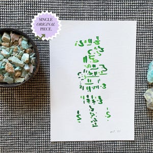 May include: A white paper with green script and the text "SINGLE ORIGINAL PIECE" in a purple circle. A bowl of turquoise and tan stones sits to the left, with two stones to the right. The background is a black and white checkered pattern.