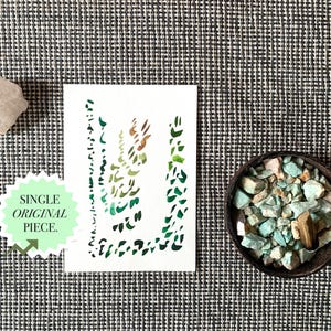 May include: A white card with a green and brown watercolor design, a clear quartz crystal, and a small bowl filled with turquoise stones. The card has the text "SINGLE ORIGINAL PIECE."