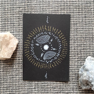May include: A black card with a circular design in gold and white, featuring stylized text and patterns. Two crystals, one white and one blue, are placed on either side of the card. The background is a black and white checkered pattern.