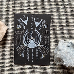 May include: A black card with white and gold geometric designs and text, flanked by two raw crystals. The card features a central circular design with radiating lines and symbols. One crystal is translucent, the other is a light blue color.