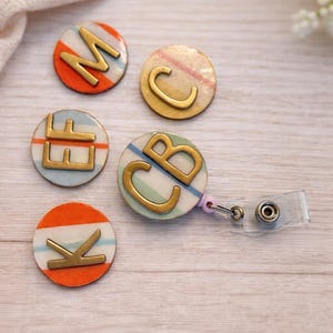 May include: A selection of personalised badge reels with gold-coloured letters. The round badges have colourful striped backgrounds in orange, blue, and green. One badge is attached to a clear retractable reel.