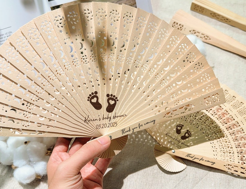 K&ouml;nnte beinhalten: Zwei Holzf&auml;cher mit einem ausgeschnittenen Design aus Sternen und Monden. Die F&auml;cher sind mit dem Text "Karen's baby shower 03.20.2025" und "Vielen Dank f&uuml;r Ihr Kommen" personalisiert.