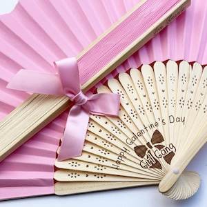 May include: Two hand fans, one with pink paper and a light wood frame, the other with a light wood frame featuring laser-engraved text reading Happy Galentine's Day Girl Gang and a bow icon. A pink satin ribbon is tied around the closed pink fan.
